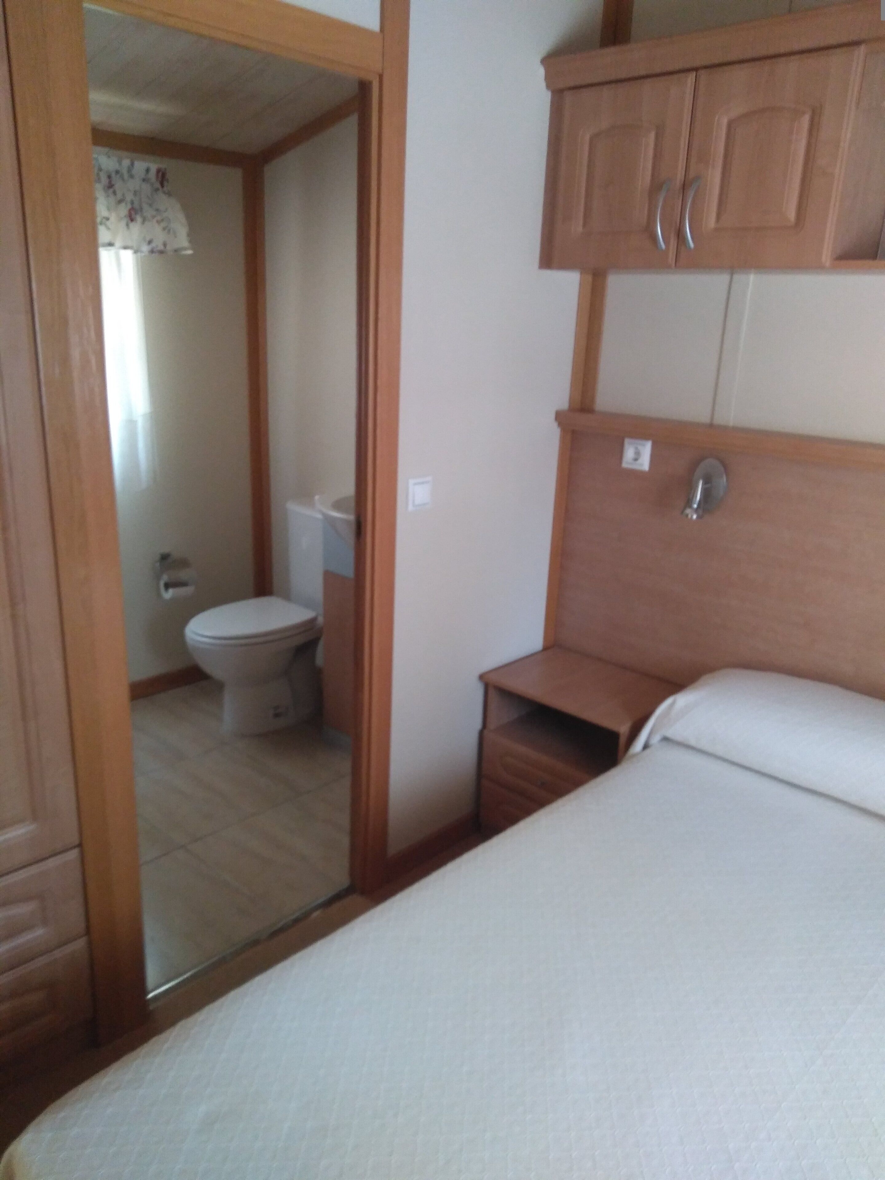 Un dormitorio con cama, armarios de madera, una mesita de noche pequeña y un baño con inodoro y lavabo.