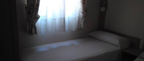 A small, dimly lit room with two single beds, a window with floral curtains, and a wooden headboard.