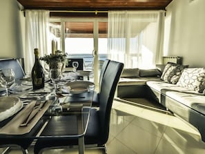 Uma sala de jantar com mesa posta para dois, um sofá e vista para o mar.