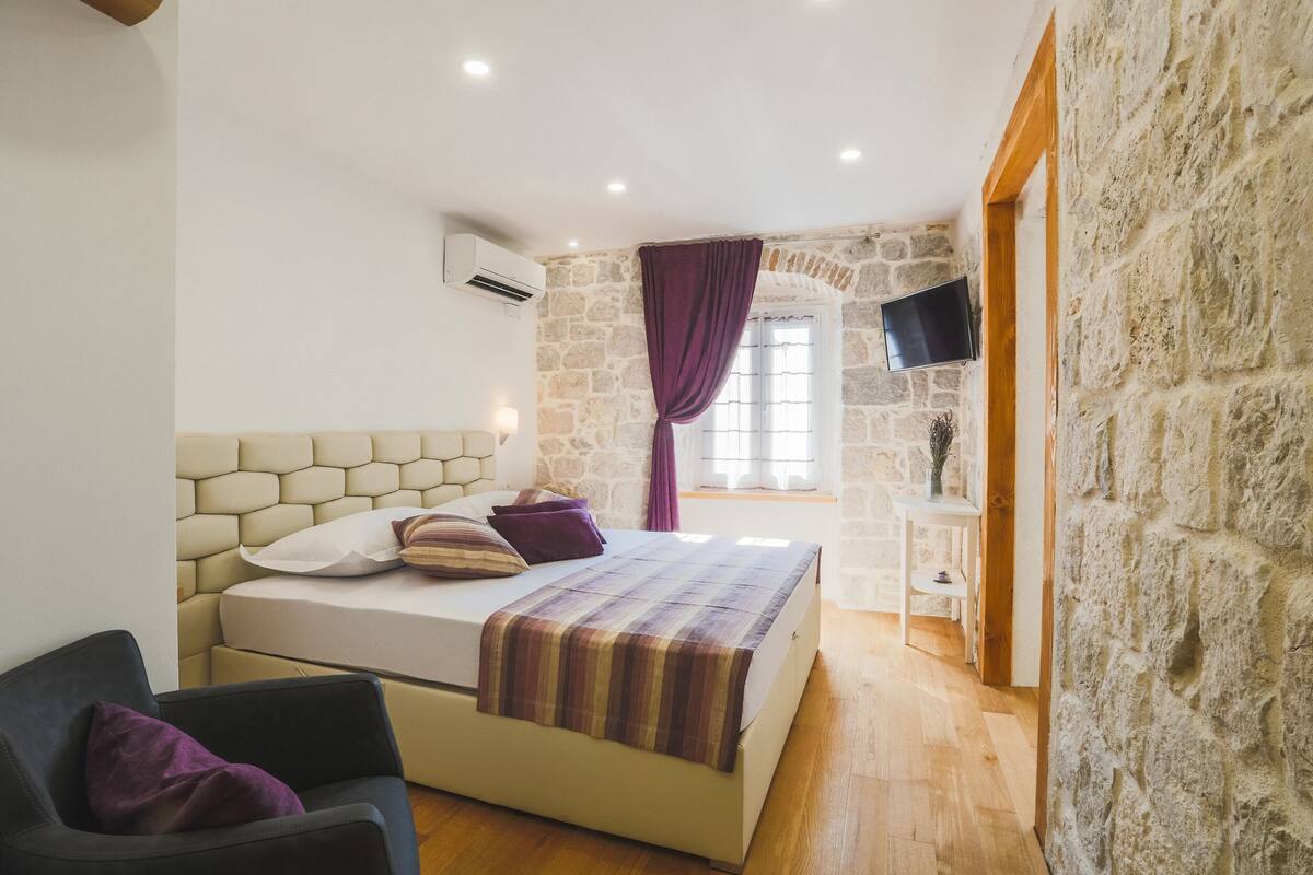 Un dormitorio con pared de piedra, una cama con manta a rayas, una silla y un televisor en la pared.