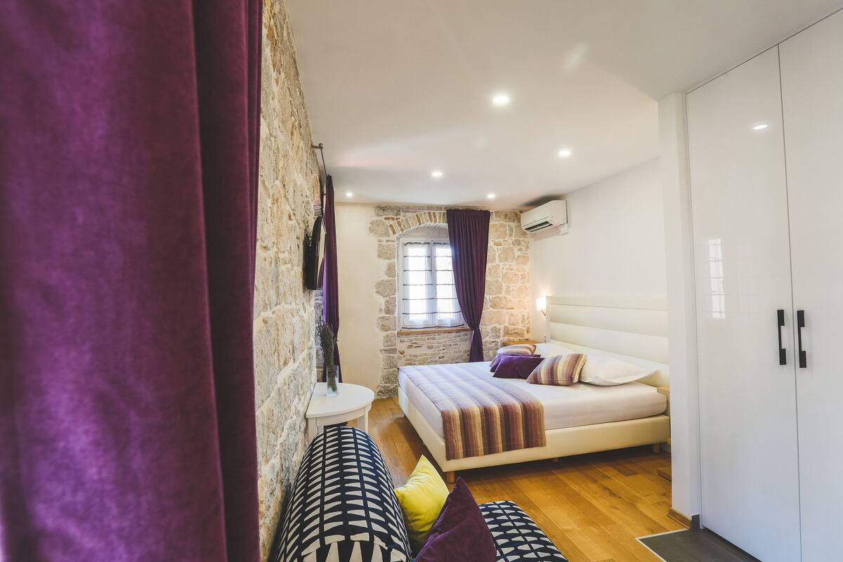 Un dormitorio con cama, armario, una mesita y ventana con cortinas.