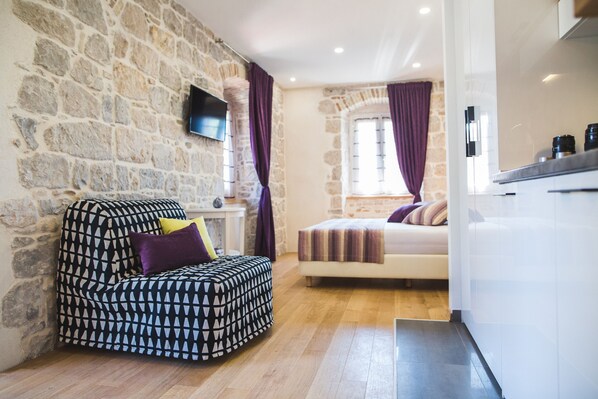 A room with a stone wall, a bed, a sofa, and a TV mounted on the wall. - Sir George Luxury Suites (Split)