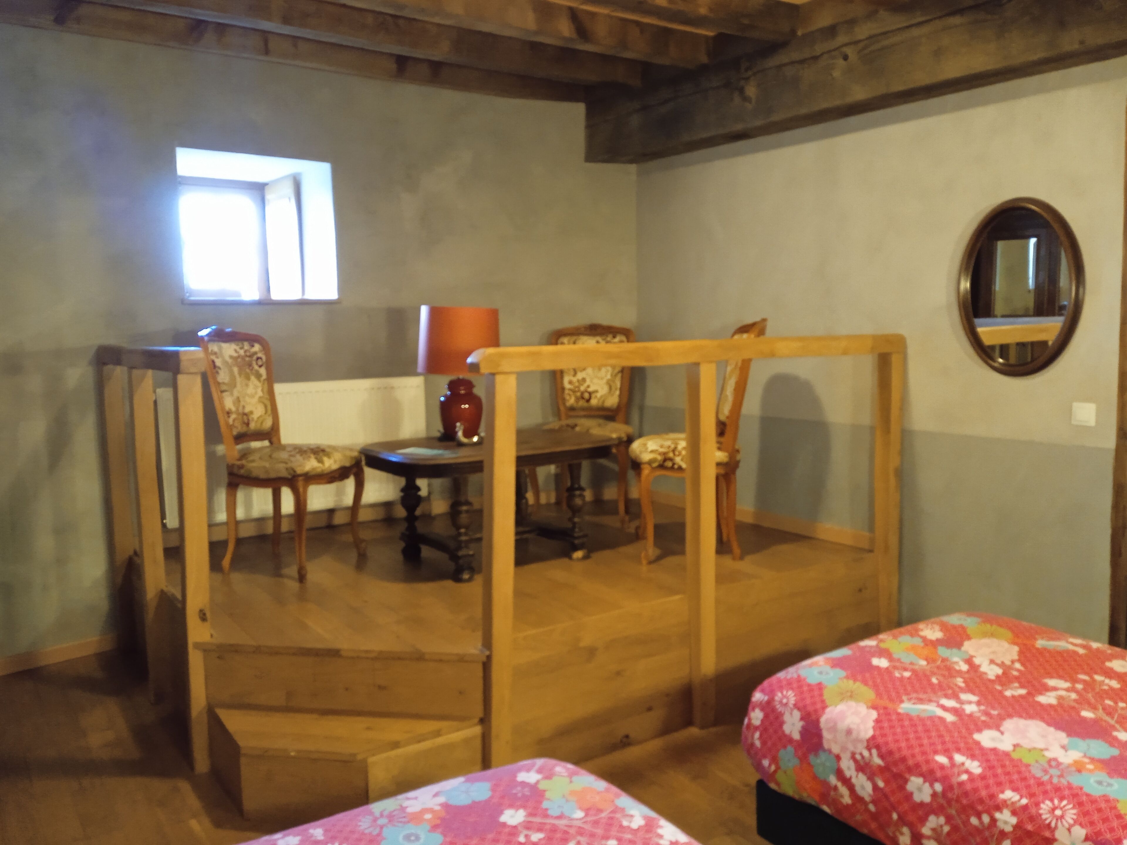 Habitació amb escala de fusta, taula de dinar amb caires, taula rodona petita amb una làmpada i un llit amb col·cha estampada de flors.
