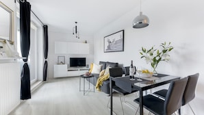 A modern living room with a dining area, white brick walls, a black sofa, a dining table with chairs, a TV, and a hanging light fixture. - P&O Apartments Metro Wilanowska 4 (Warsaw)