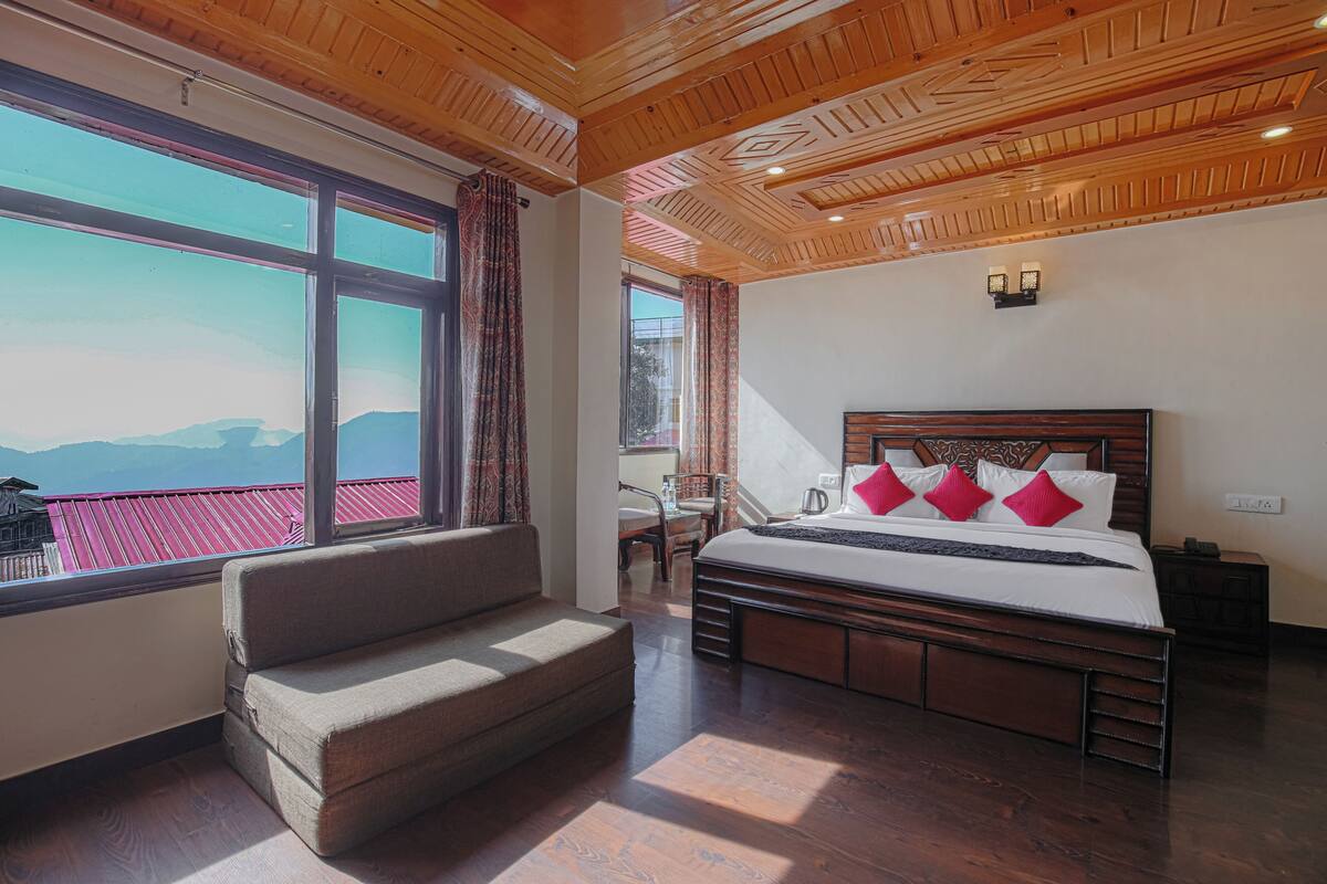 A bedroom with a large window, a bed with red pillows, a wooden ceiling, and a view of mountains.