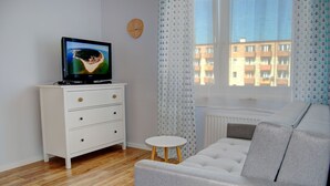 En moderne stue med et fladskærms TV på en hvid kommode, en grå sofa, et lille rundt bord og udsigt til en bygning gennem lette gardiner.