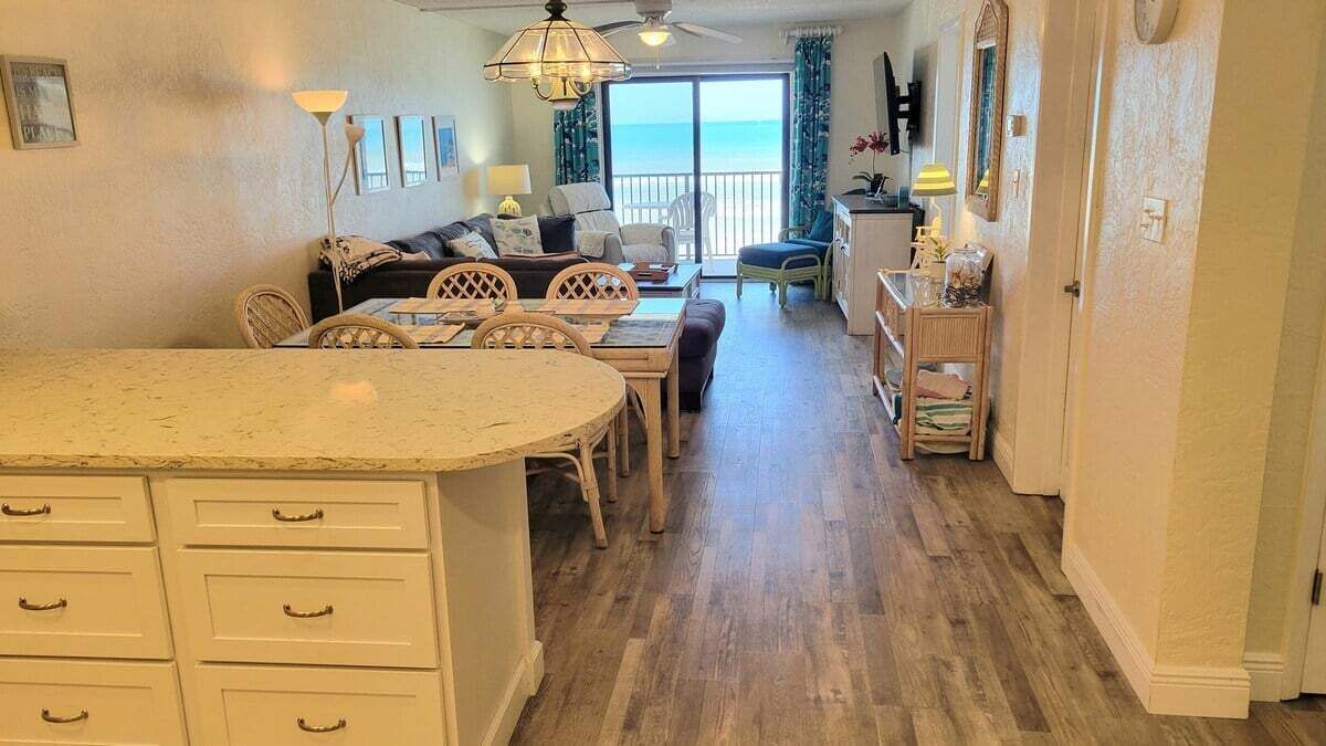A spacious living area with a large island, dining table, and chairs, leading to a balcony with a view of the ocean.