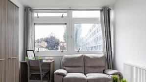 En moderne stue med en grå sofa, et glasbord, et træskrivebord og udsigt til bygninger gennem store vinduer.