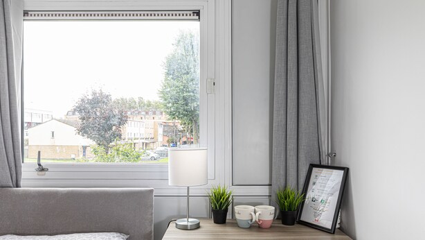 Et soveværelse med en seng, et skrivebord med lampe, to potteplanter og en rammet menu på væggen.