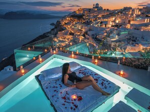 A woman relaxing on a bed with rose petals, overlooking a coastal town with white buildings and a clear view of the sea. - Saint Santorini (Santorini)