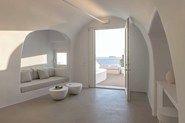A modern interior with a curved sofa, two round tables, and a view of the sea through an open door. - Saint Santorini (Santorini)
