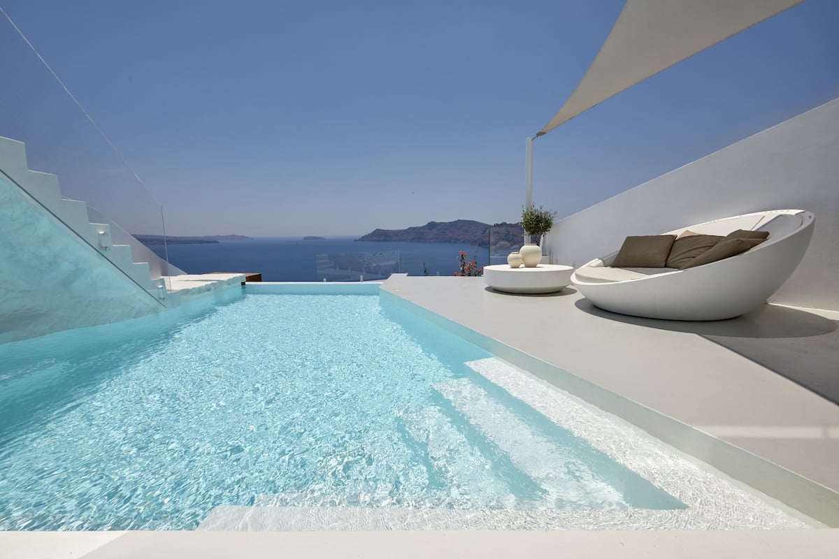 A modern infinity pool with clear blue water, a white infinity edge, and a white infinity deck. There is a white infinity chair with cushions and a small table with a plant.