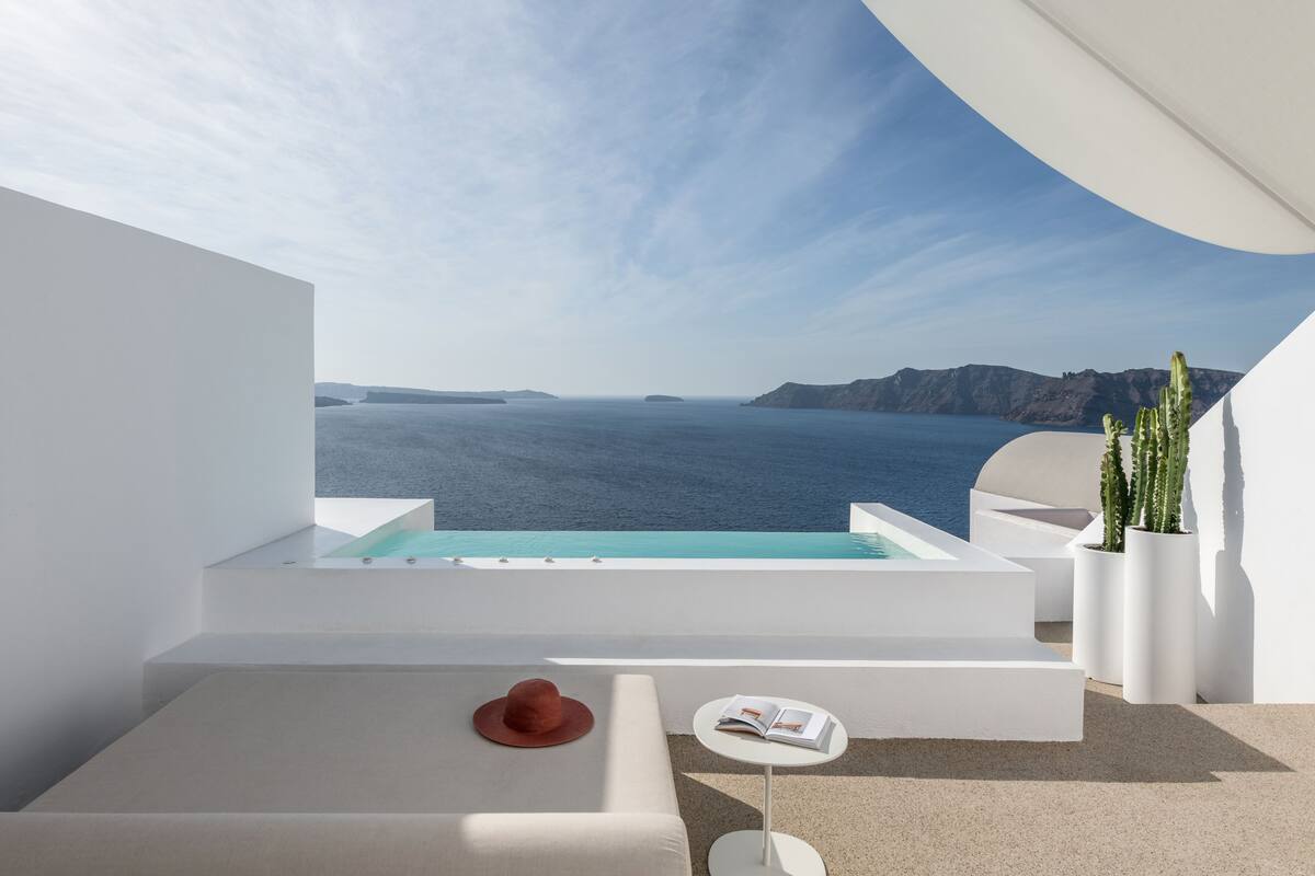 A modern balcony with a pool, white furniture, and a view of the sea and cliffs.