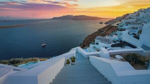 Una escalera que baja hacia una zona costera con edificios blancos, un barco en el agua y un atardecer de fondo.