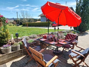 Une terrasse extérieure avec une table dressée pour le repas, entourée de chaises en osier et d’un parasol rouge.