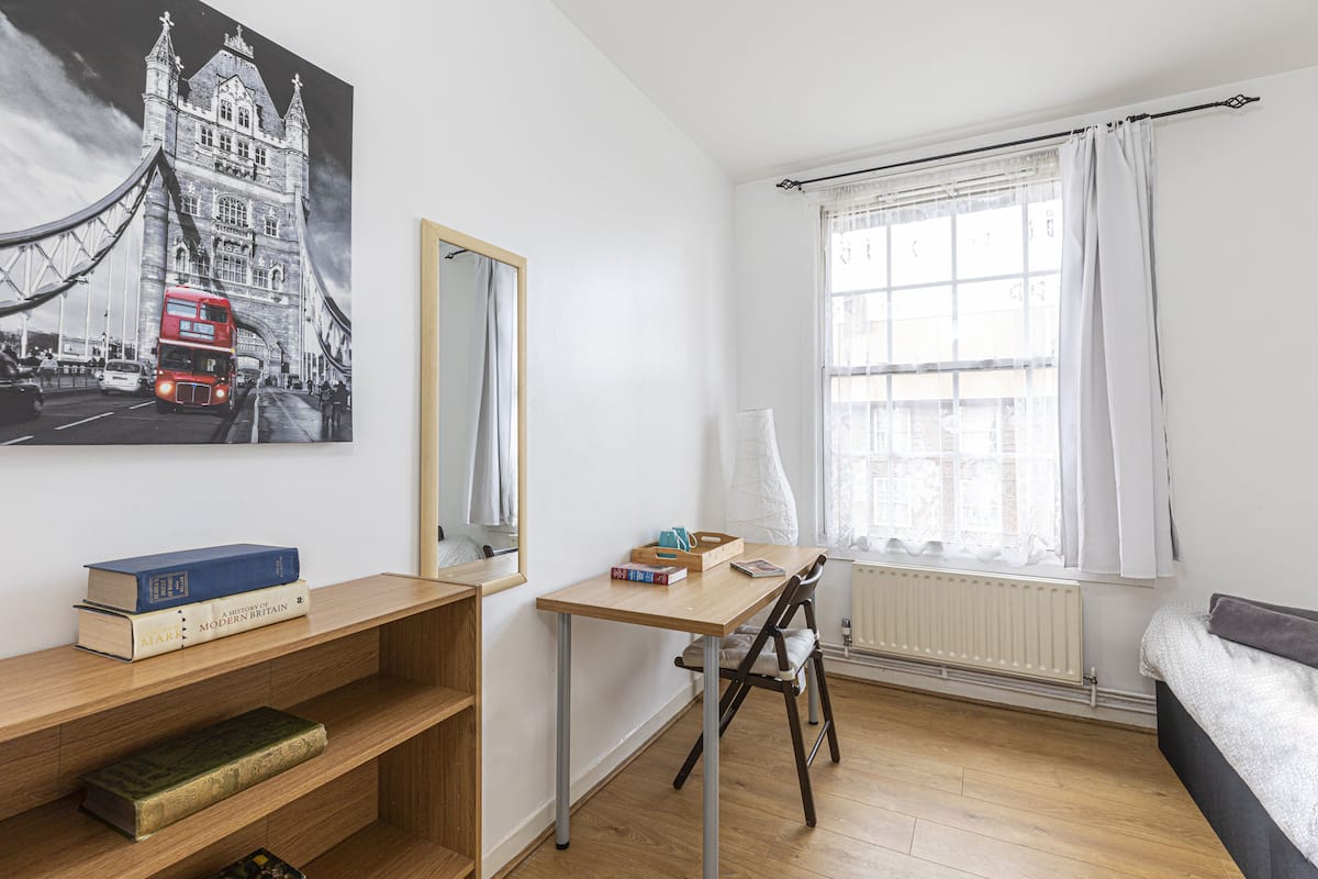 A room with a wooden desk, a chair, a bookshelf with books, a large window with curtains, and a wall-mounted mirror.