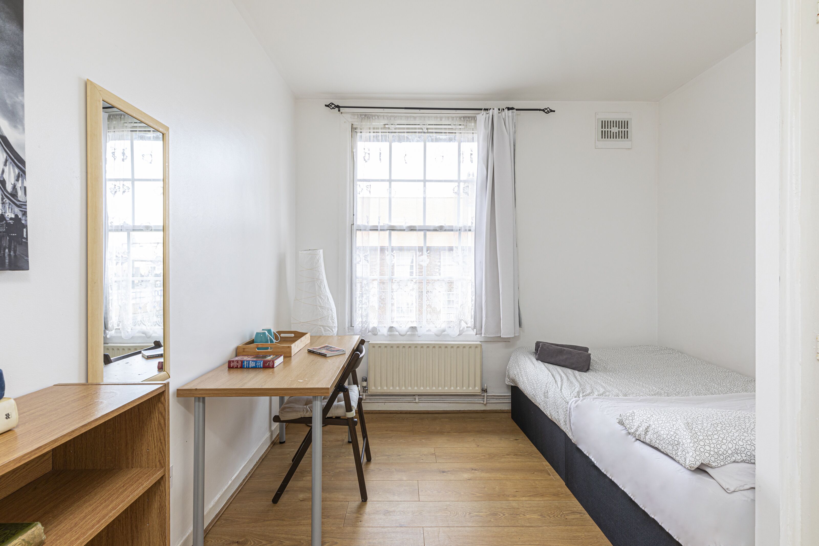 A small, well-lit room with a wooden desk, a bed with a white and gray patterned bedspread, a radiator, and a window with white curtains.