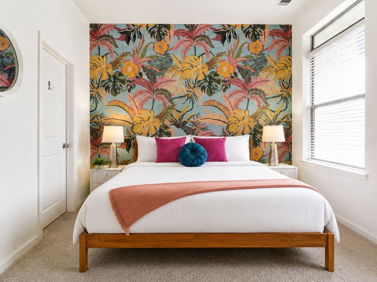 A bedroom with a bed, two bedside lamps, a wall mural, and a window with blinds.