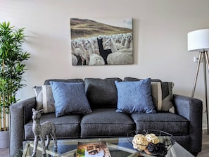 A living room with a grey sofa, blue cushions, a glass coffee table with decorative items, a wall-mounted painting of llamas, and a bamboo plant.