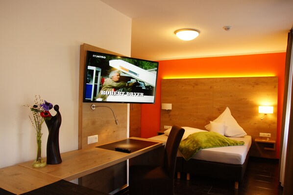 Habitación de hotel con cama, escritorio con lavamanos, televisor montado en la pared y cabecero de madera.