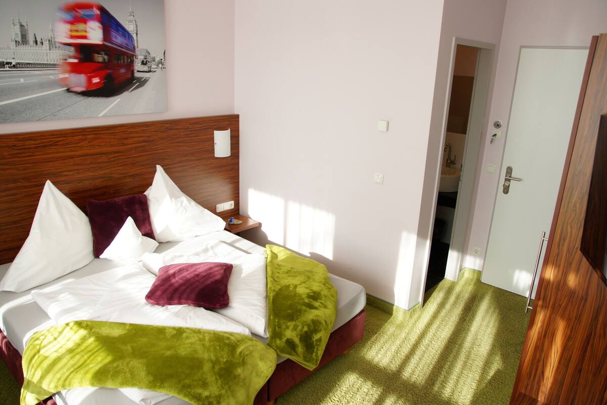 Una habitación de hotel con una cama, una mesita de noche, un cuadro de un autobús rojo de dos pisos y una puerta que da a un baño.