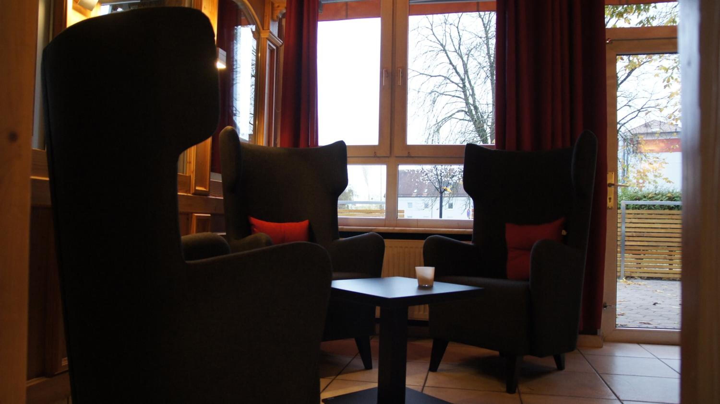 A cozy seating area with two armchairs and a small table, large windows with red curtains, and a view of the outside.