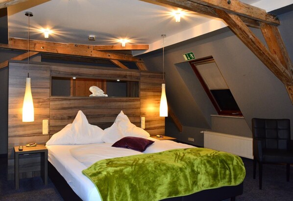 Un dormitorio con techo estilo loft, vigas de madera, una cama con ropa blanca, una manta verde y dos lámparas de noche.