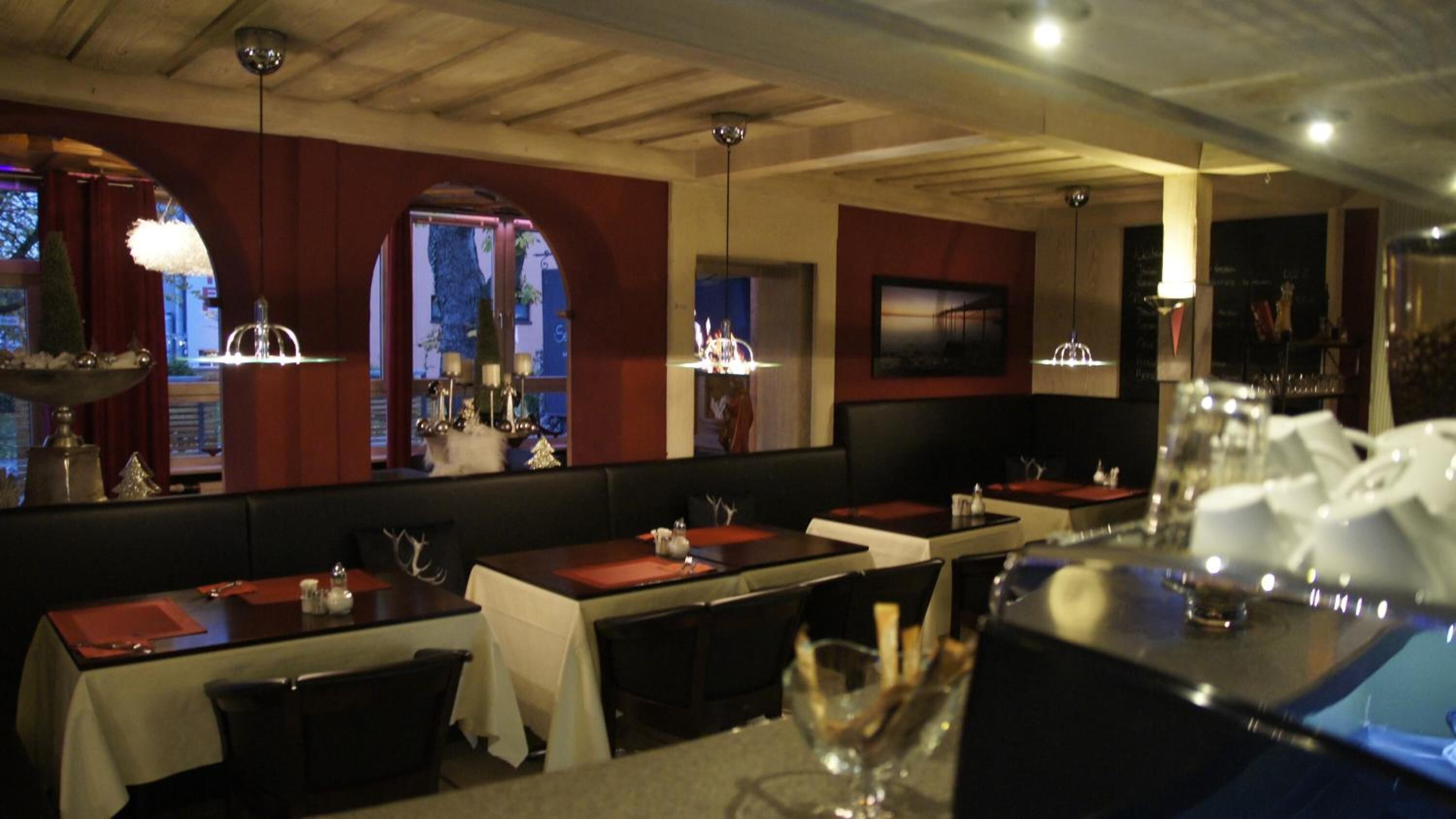 Un restaurante con varias mesas, cada una con mantel blanco y posavasos rojos. Luces de araña cuelgan del techo y las paredes están decoradas con cuadros enmarcados.