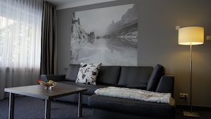Una habitación de hotel moderna con un sofá gris, una pequeña mesa de centro de madera, una lámpara y un cuadro abstracto grande en la pared.