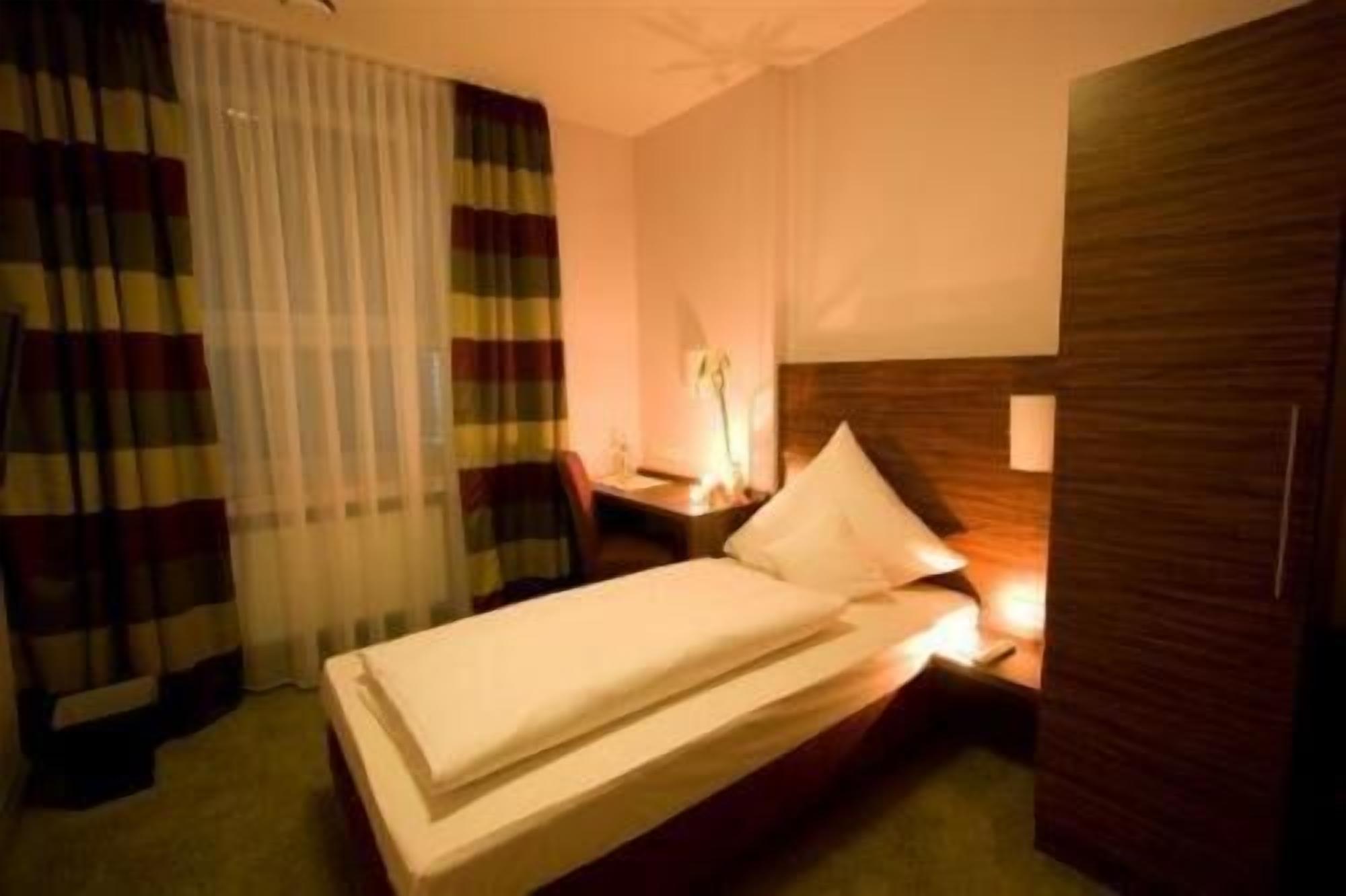Habitación de hotel con una cama individual, una mesita de noche con lámpara, una silla y un armario de madera.