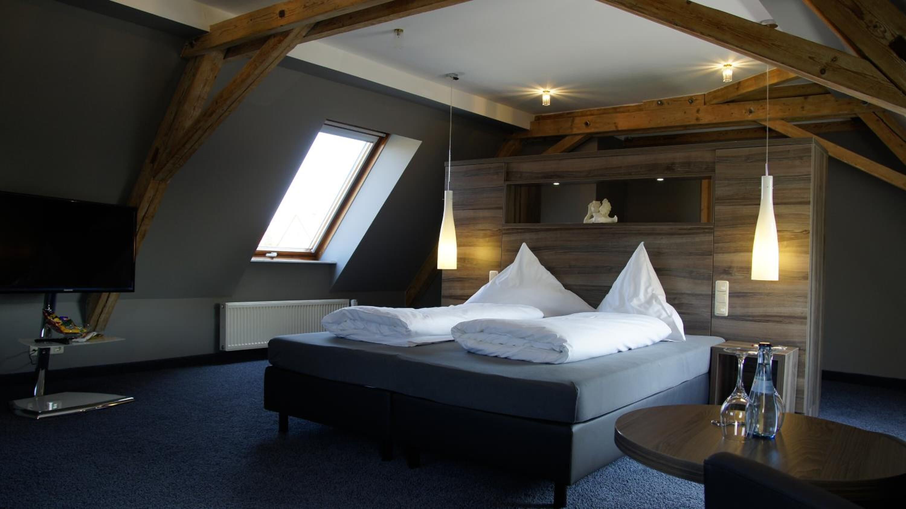 Un dormitorio moderno con techo inclinado, una cama grande con ropa de cama blanca, un televisor y una mesita con vasos.