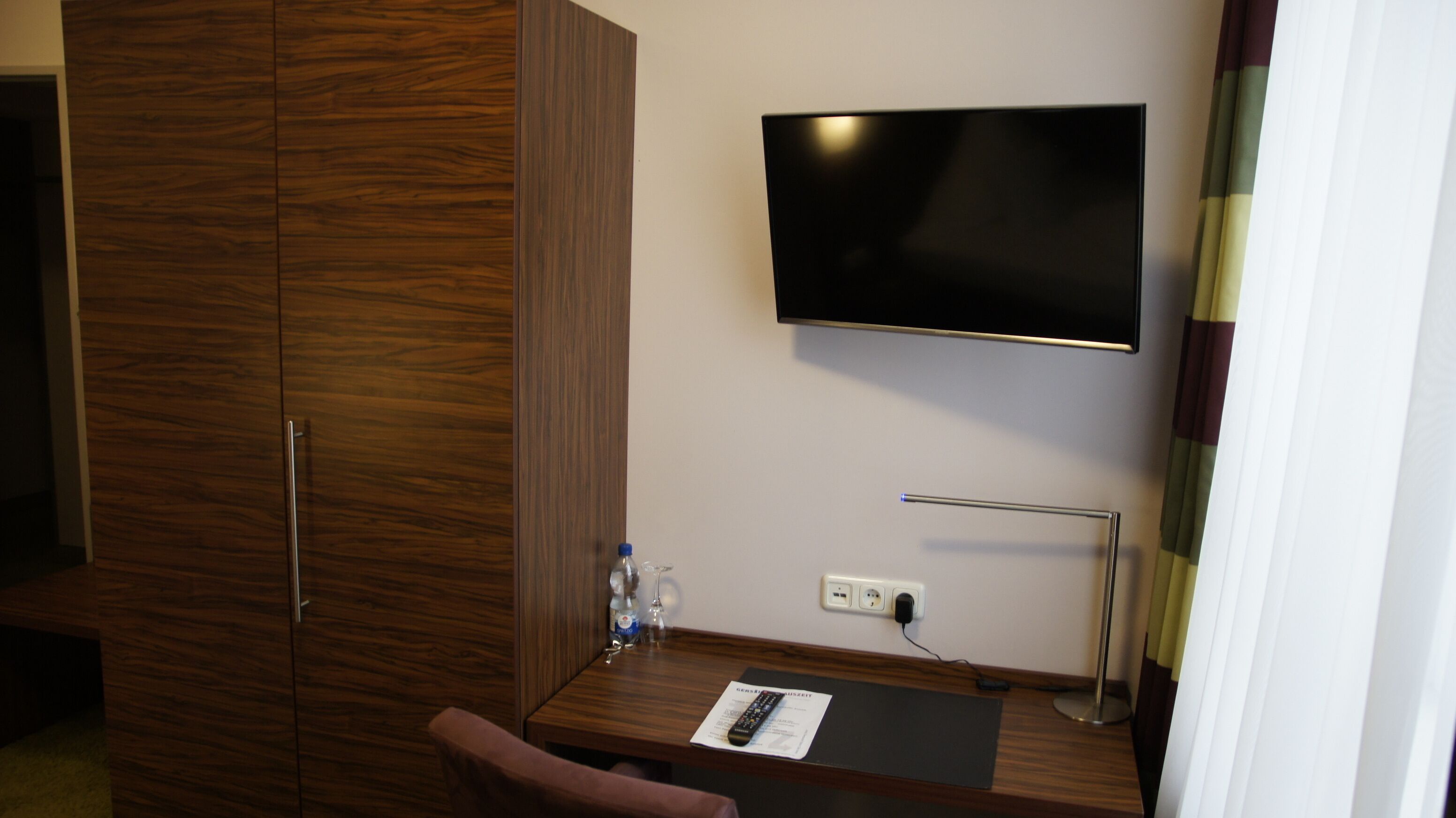 Une chambre d’hôtel équipée d’une armoire en bois, d’un téléviseur à écran plat fixé au mur, d’un bureau avec une télécommande, d’une bouteille d’eau et d’un verre.