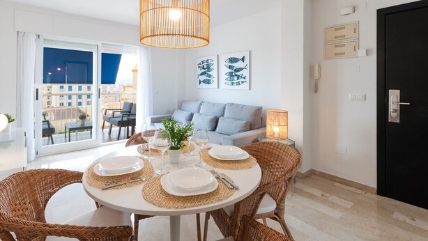 Un coin repas avec une table ronde dressée pour quatre personnes, entourée de chaises en osier, d’un luminaire suspendu et d’un canapé en arrière-plan.