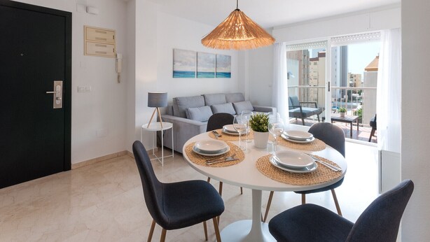 Uma área de jantar moderna com uma mesa redonda posta para quatro pessoas, uma luminária pendente e vista para a cidade através de grandes janelas.