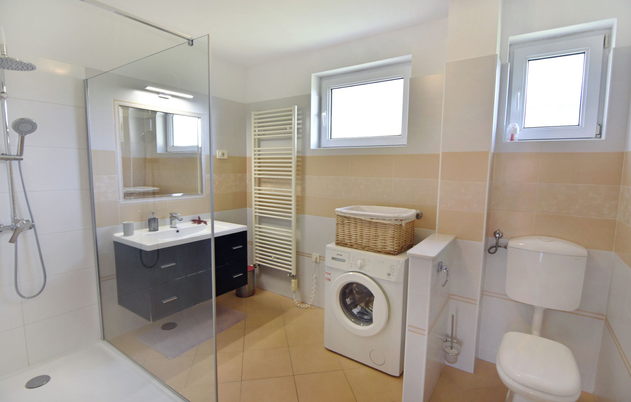 A bathroom with a glass-enclosed shower, a washing machine, a toilet, and a window above the sink.