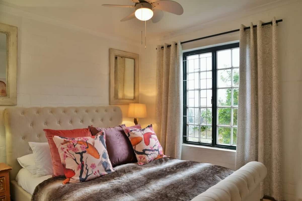 A bedroom with a tufted headboard, a large window with curtains, and a bed with patterned pillows.
