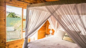A wooden cabin bedroom with a bed under a canopy, a nightstand with a lamp, and a view of a fenced garden. - Anna Beulah Farm (Cape Town)
