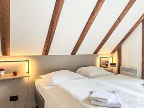 Ein Schlafzimmer mit geneigter Decke, Holzbalken, einem Bett mit weißer Bettwäsche, einem Nachttisch mit Lampe und einem Wandheizkörper.