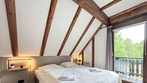 A bedroom with a large bed, wooden beams, a nightstand with a lamp, and a view of greenery outside.
