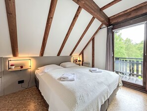 Ein Schlafzimmer mit einem großen Bett, Holzbalken, einem Nachttisch mit Lampe und Blick auf Grünflächen.