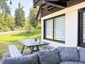 A patio with a table and a sofa, a bottle of champagne, and a car parked on the road in the background.