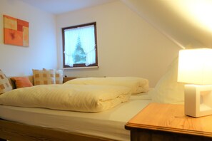 Un dormitorio con cama, almohadas, una mesita de noche, lámpara y una ventana con cortinas.