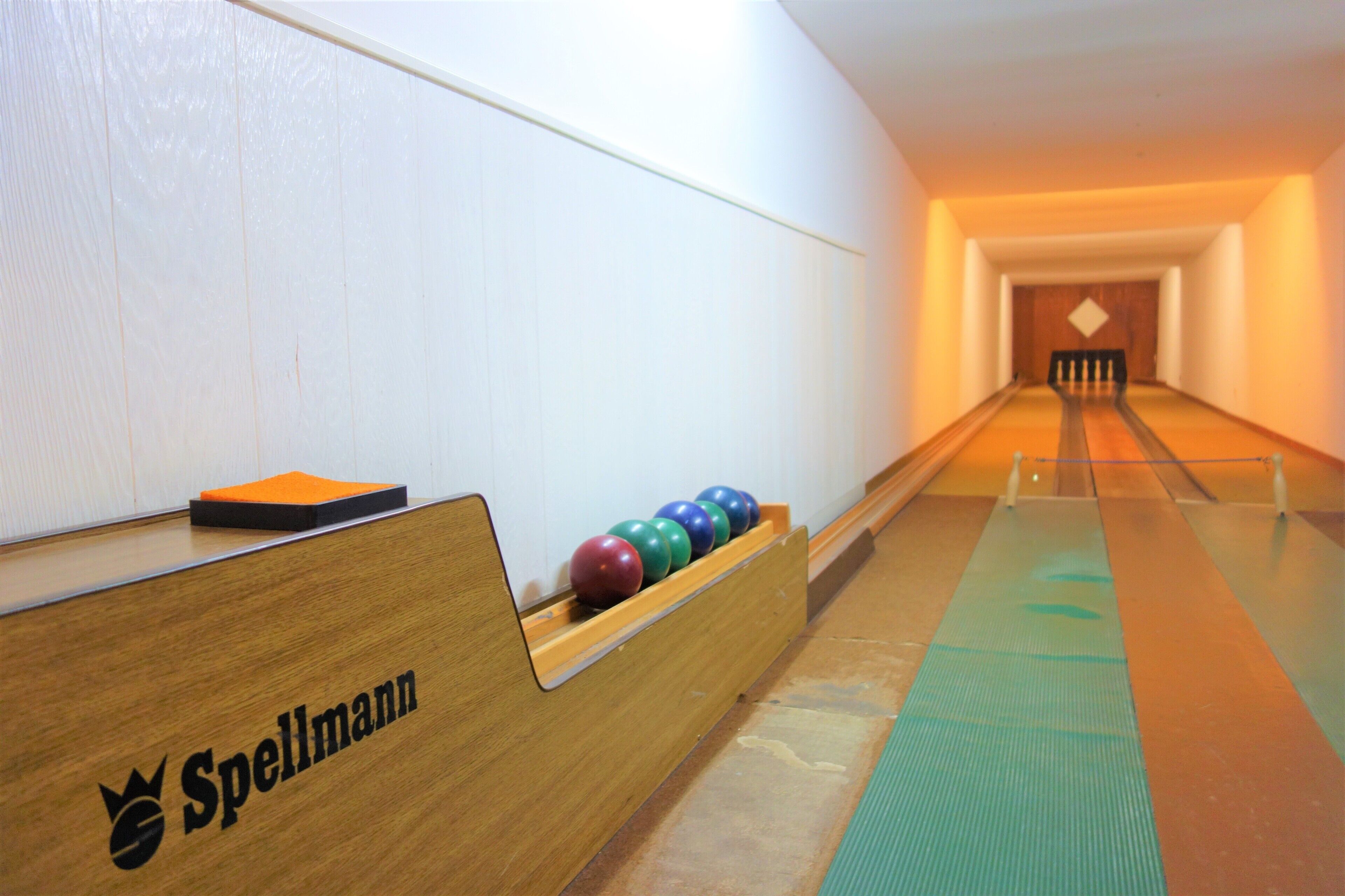 A bowling alley with a wooden counter labeled "Spellmann" and a row of colorful bowling balls.