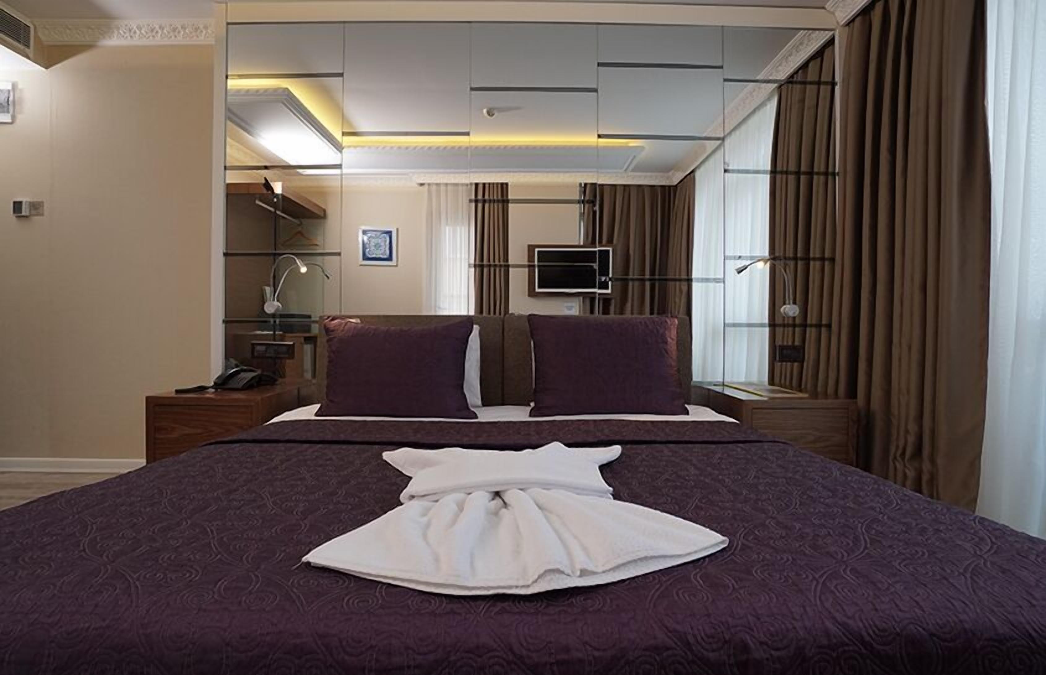 A hotel room with a large bed, two pillows, a bedside table with a lamp, a TV, and a window with curtains.