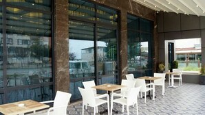 An outdoor café with white chairs and tables, large windows, and a tiled floor. - Otel 1919 (Atakum)