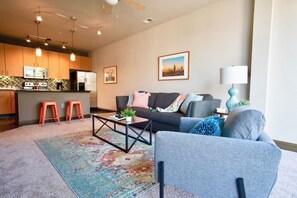 A modern living room with a sofa, coffee table, and a rug. There is a kitchen area with a microwave and stools.