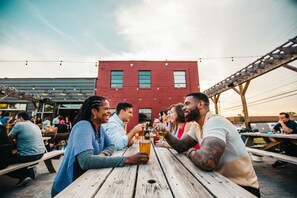 A group of people seated at wooden picnic tables outdoors, with a red brick building in the background.