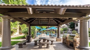 Eine Terrassenanlage im Freien mit einer Pergola, Steinpfeilern, einem Grill und einem Pool im Hintergrund.