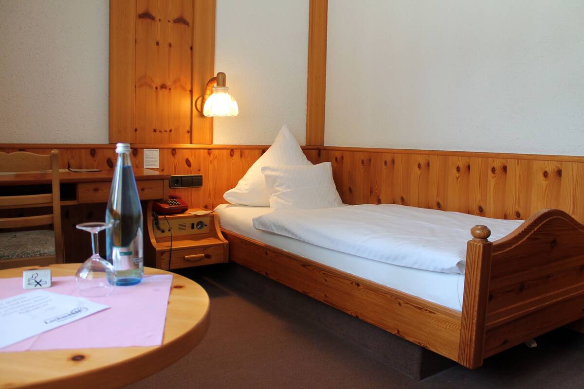 A wooden bed with white bedding, a bedside lamp, a wooden nightstand, a chair, and a table with a bottle and glass.