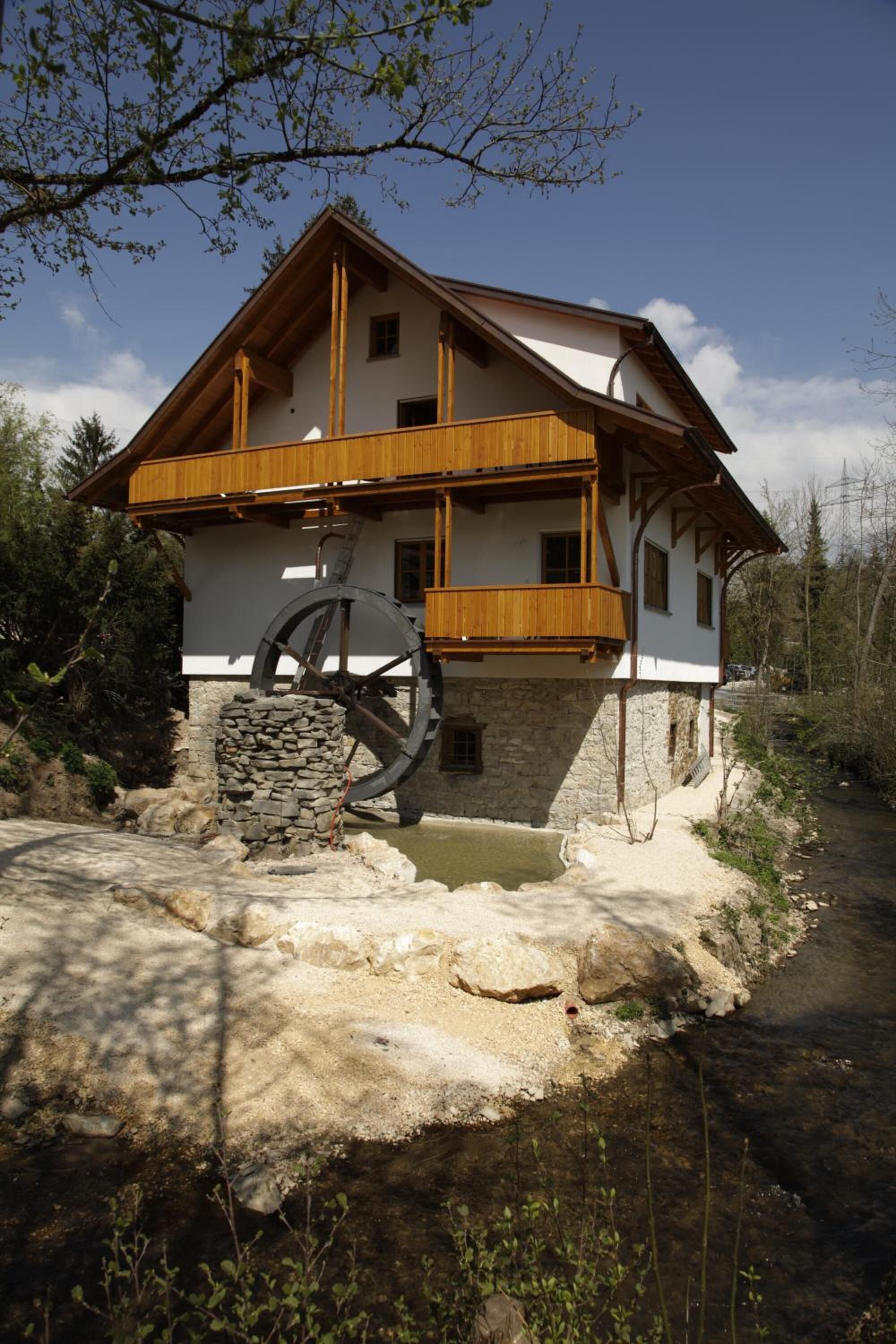 Ein zweigeschossiges Holzhaus mit Steinuntergrund, einem großen Wasserrad und einem Feldweg, der zum Eingang führt.